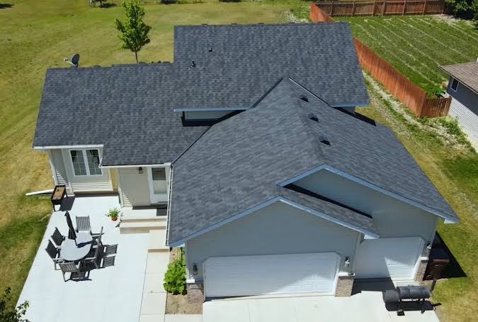 An aerial view of a single-story home with a newly installed charcoal grey architectural shingle roof, light grey siding and white trim, a concrete patio with outdoor furniture, and a wooden privacy fence bordering the lawn
